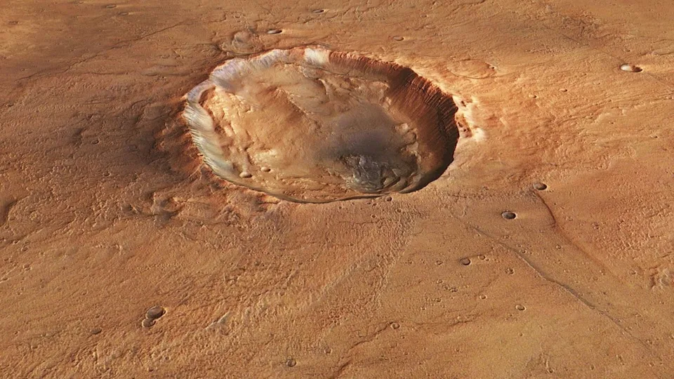 A photo of the surface of Mars from space, with a large crater seen in its reddish brownish surface.