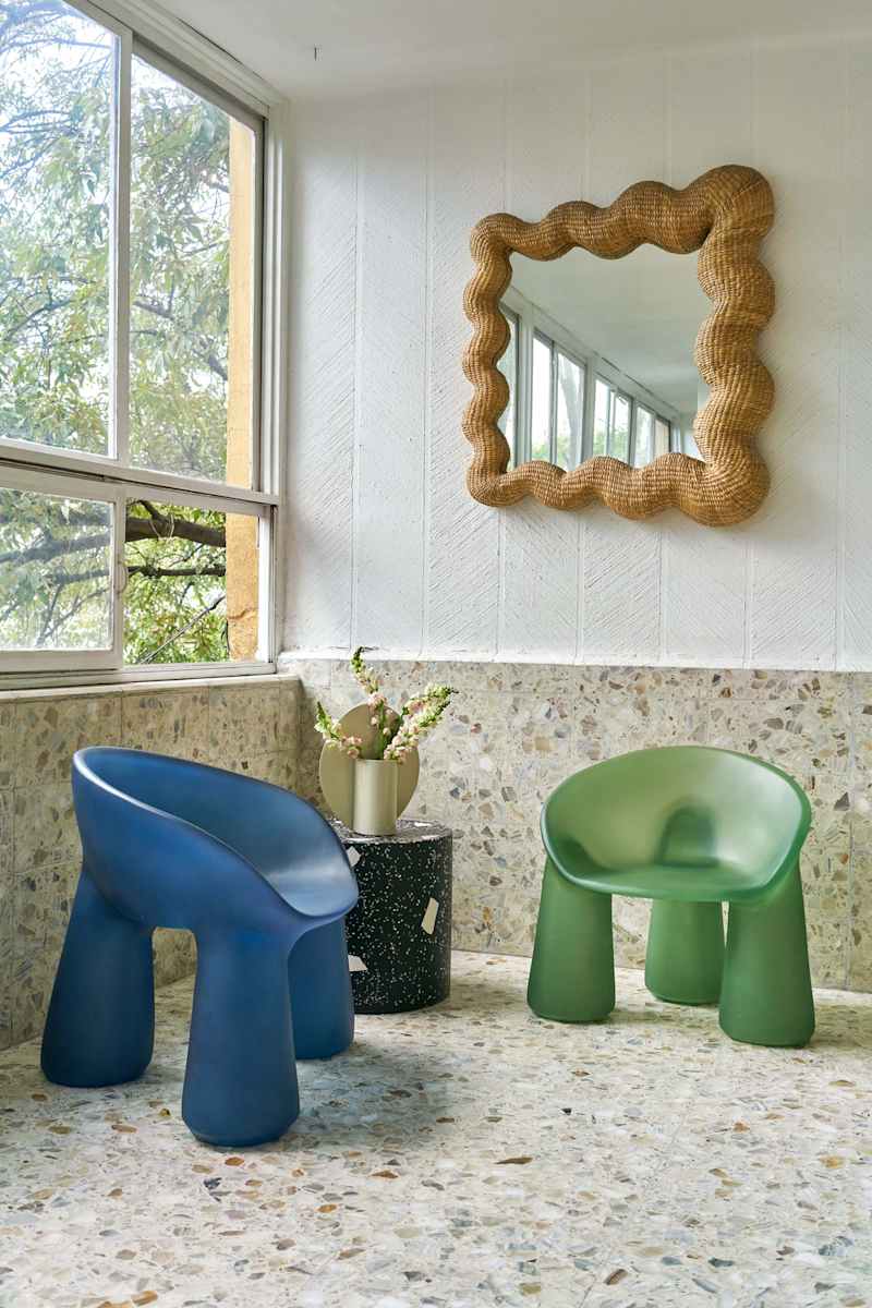 Two colorful chairs, one blue and one green, beside a black speckled table with a vase of flowers, and a wavy mirror on the wall.