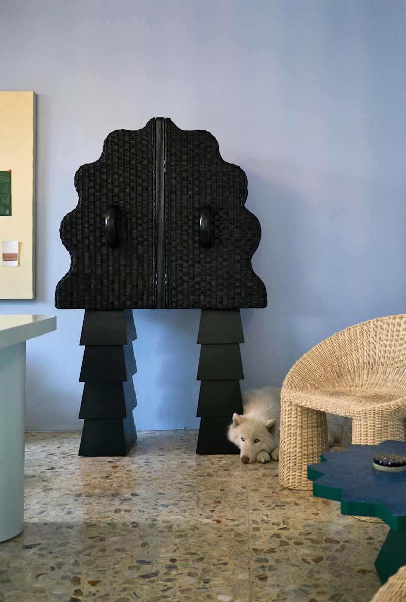 A black cabinet with a scalloped top stands on tiered legs, next to a woven chair and a white dog resting on the floor.