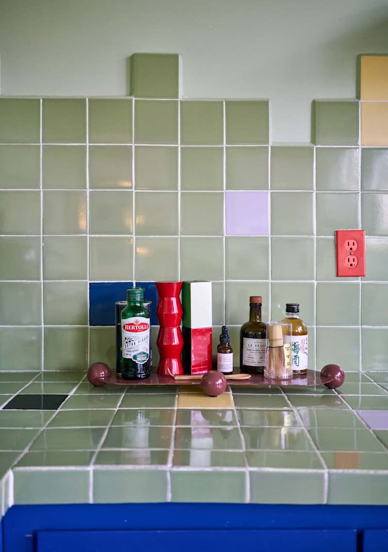 Colorful kitchen countertop with green tiles, various bottles, and decorative items, including a red vase and wooden spoons.