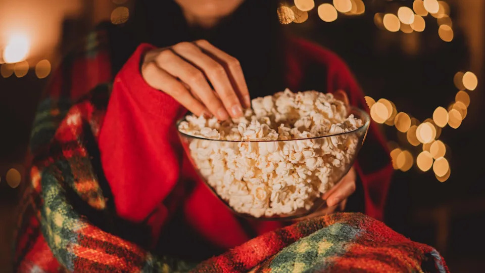 Cheerful woman eating popcorn and watches christmas comedy movie on cable TV at home in evening alone, close-up. Christmas traditions concept