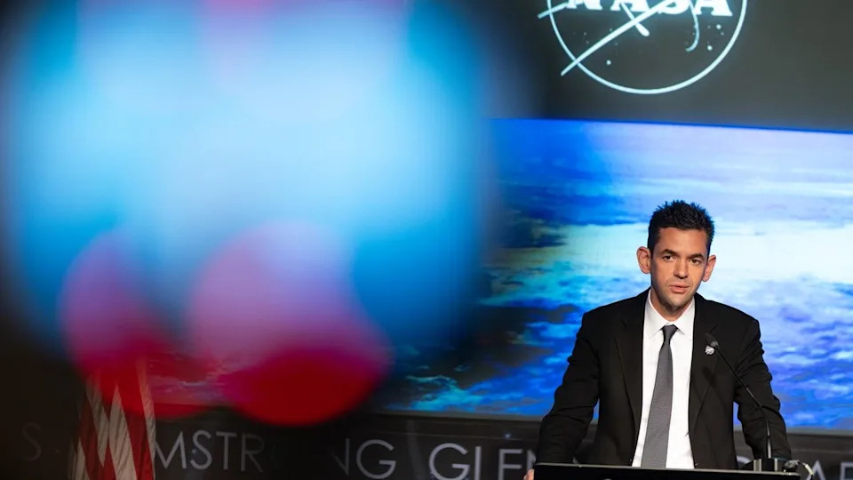 A man in a dark suit speaks at a lectern in front of an image of earth on a large screen behind him.